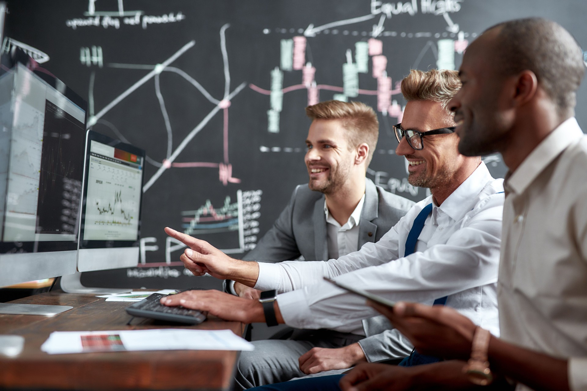 Dreams Come True. Side view of three colleagues traders sitting by desks in front of computer monitors and discussing strategies. Blackboard full of charts and data analyses in background